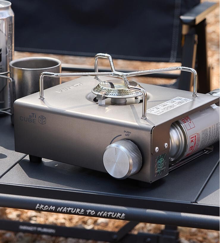 The Kovea Titan Cube camping burner set up on a wooden table at an outdoor campsite, blending in with nature.