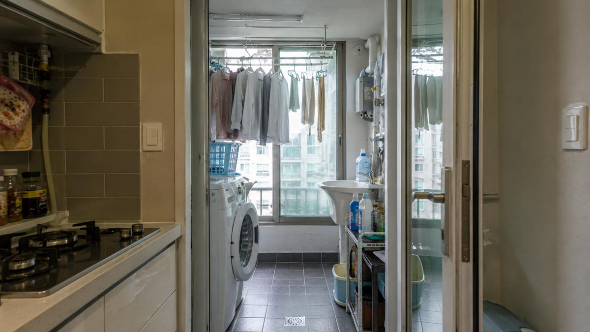 A separate Korean apartment utility balcony with a washing machine, ceiling drying rack, utility sink, and household supplies.