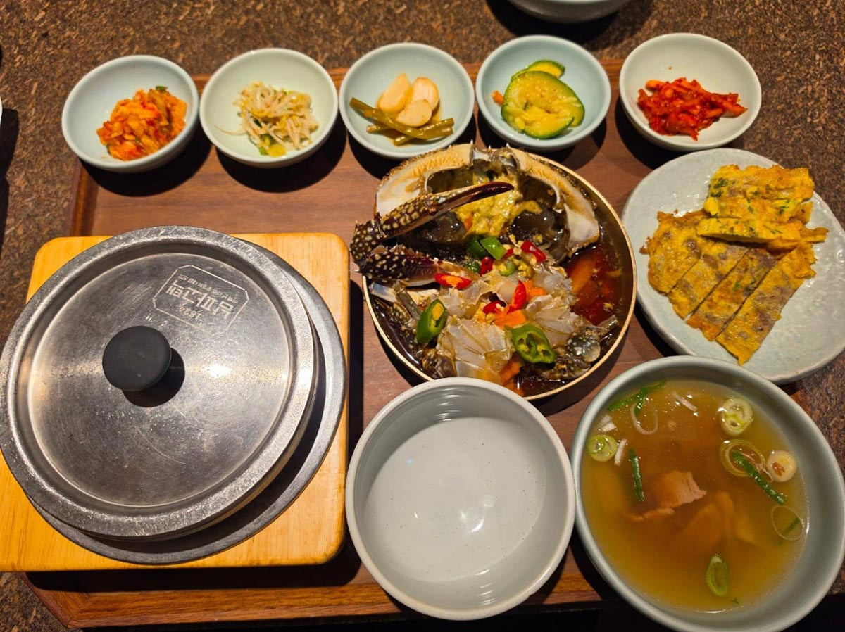 Korean banchan meal featuring marinated crab, kimchi, seasoned vegetables, jeon, rice, and soup