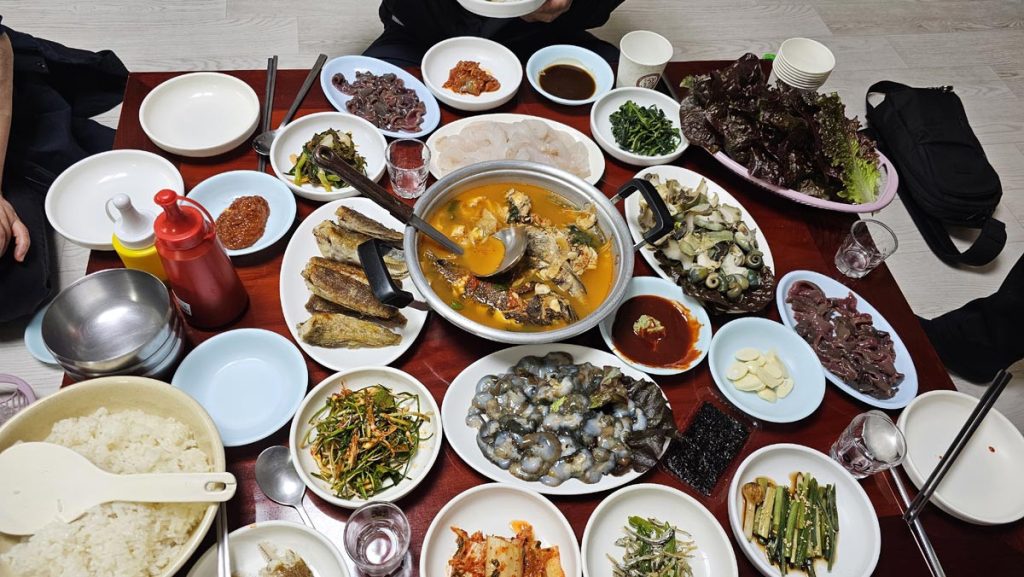 Korean shared dining table with banchan, seafood, fish stew, raw octopus, vegetables, rice, and seaweed