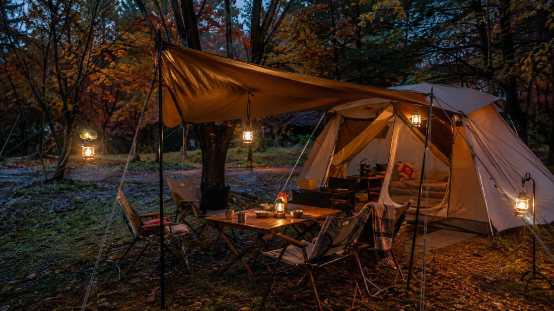 Korean campers' outdoor living room with warm lanterns, wooden table, chairs, and a tent in an autumn forest campsite