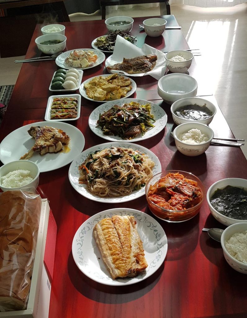 Korean home meal served with rice, soup, japchae, kimchi, grilled fish, and several homemade banchan