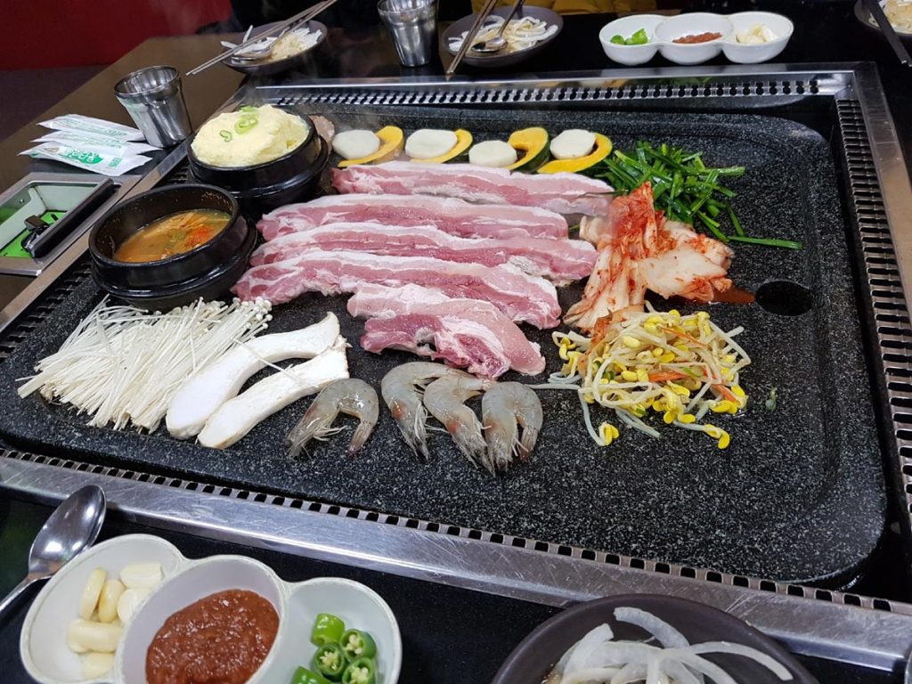 Korean samgyeopsal BBQ with thick pork belly, kimchi, vegetables, mushrooms, and shrimp ready to grill at a restaurant