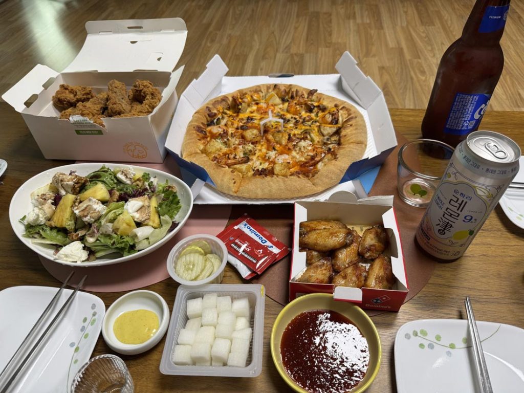 Korean-style delivery spread with fried chicken, pizza, chicken wings, salad, pickled radish, and dipping sauces