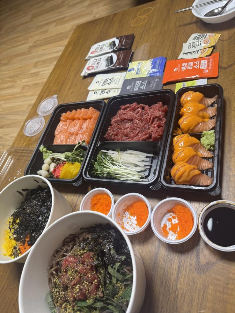 Korean delivery meal with salmon sashimi, spicy salmon sushi, yukhoe, rice bowls, and dipping sauces