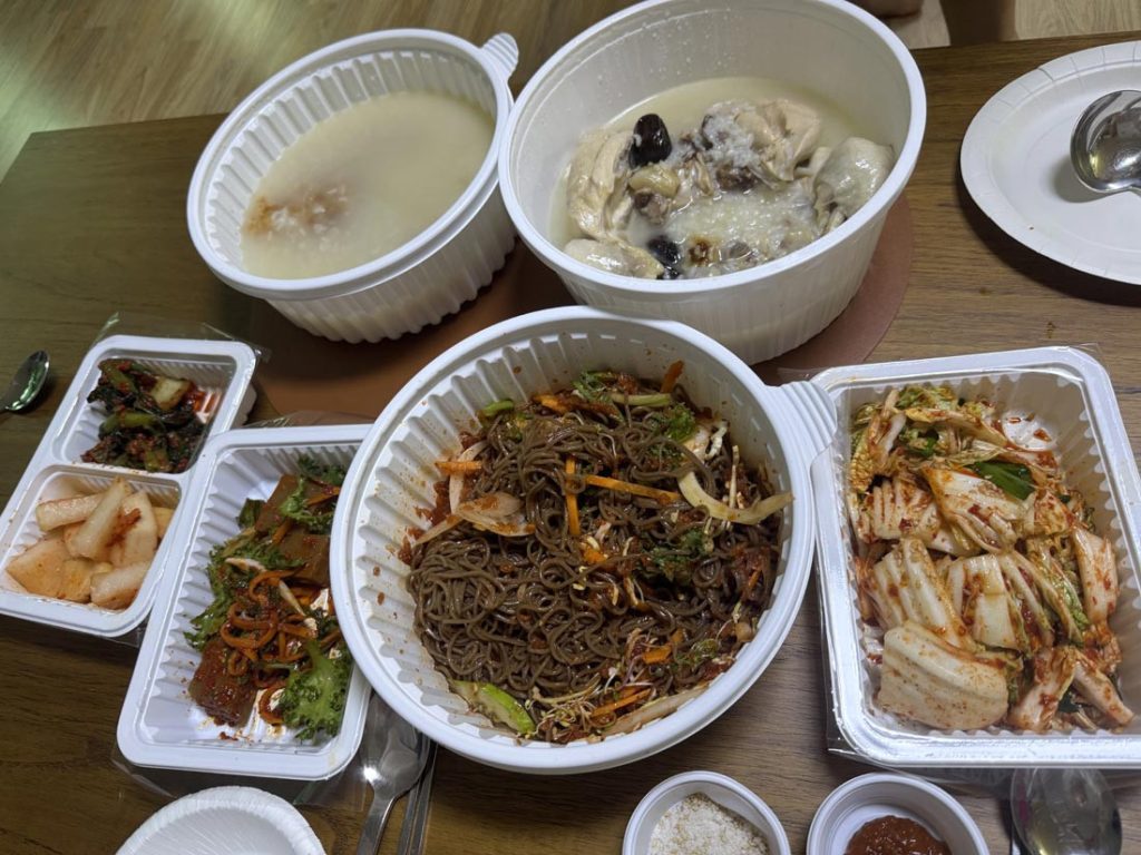 Korean delivery meal with samgyetang chicken soup, spicy makguksu noodles, fresh kimchi, and side dishes